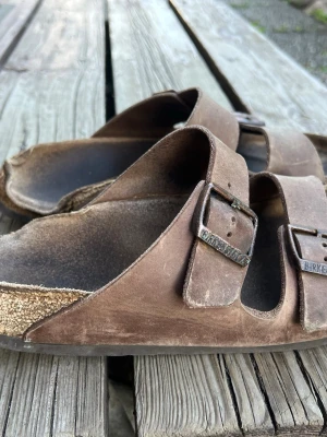 Bruna Birkenstock sandaler i mocka - Klassiska bruna sandaler från Birkenstock med två justerbara remmar och metallspännen. Ovandelen är i mocka och sulan är formad för extra komfort. Perfekta för en avslappnad stil och passar till det mesta.