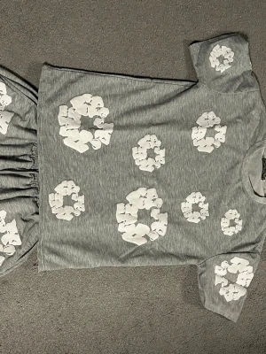 Grå t-shirt från Denim Tears - Grå t-shirt från Denim Tears med vita blommiga tryck över hela tröjan. T-shirten har rund halsringning och korta ärmar. Perfekt för dig som vill ha en unik och trendig topp med coolt mönster.