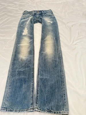 Blå slitna bootcut jeans - Säljer ett par blå bootcut jeans med slitningar och distressed detaljer på låren och knäna. Jeansen har klassisk femficksmodell, låg midja och ljusa tvättade partier. Perfekta för en avslappnad och trendig look.