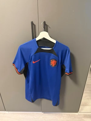 Blå Nederländerna Nike fotbollströja - Snygg blå fotbollströja från Nike med Nederländernas landslagsemblem på bröstet och orange Nike-logga. Tröjan har svarta detaljer vid axlar och sidor samt orange kant vid ärmslut. Perfekt för fotbollsträning eller match.