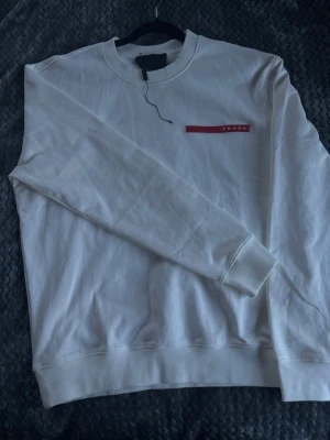 Vit sweatshirt från Prada - Snygg vit sweatshirt från Prada med rund halsringning och diskret röd logga på bröstet. Tröjan har långa ärmar, ribbade muddar och är tillverkad i mjukt material som känns skönt mot huden. Perfekt för en clean och stilren look. Vid frågor kontakta mig via dm svara så snabbt jag kan 