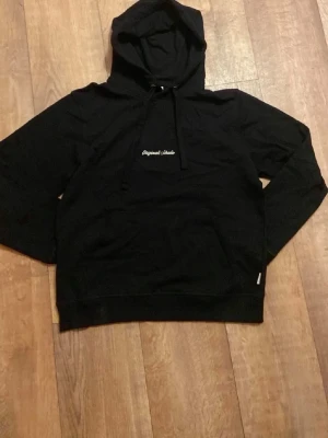 Svart hoodie från Jack n Jones  - Svart hoodie från Original Classic med broderad vit text på bröstet. Klassisk modell med huva och dragsnören, samt ribbade muddar vid ärmslut och nederkant. Perfekt för en avslappnad och stilren look.