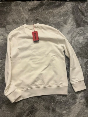 Vit sweatshirt från Hugo, storlek S - Säljer en stilren vit sweatshirt från Hugo i mjuk bomull. Tröjan har klassisk rund hals, ribbade muddar och en clean look utan tryck på baksidan. Perfekt för dig som gillar minimalistisk och modern stil.