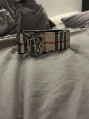 Burberry bälte  - Snyggt bälte från Burberry med klassiskt rutmönster i beige, svart och rött. Bältet har ett stort silverfärgat B-spänne och är gjort i skinn. Perfekt accessoar för att lyfta din outfit med en lyxig touch.