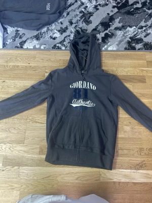 Mörkgrå hoodie från Giordano - Mörkgrå hoodie med dragkedja från Giordano. Tryck på bröstet med texten 'Giordano 81 Authentic' i vitt och blått. Utrustad med huva och två fickor framtill. Perfekt för en avslappnad streetwear-look.