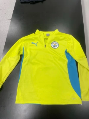 Manchester City träningströja gul - Gul långärmad träningströja från Puma med Manchester City-logga på bröstet och blå detaljer längs sidorna. Tröjan har halv dragkedja framtill och är tillverkad i ett lätt, ventilerande material. 10-14 år passar som xs-s 