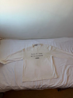 Vit långärmad Acne Studios t-shirt - Vit långärmad t-shirt från Acne Studios med tryck på bröstet där det står 'Acne Studios STOCKHOLM 1996'. Klassisk rund hals och relaxed fit. Perfekt för dig som gillar minimalistisk och stilren design.