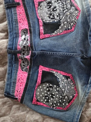 Jeansshorts med dödskallepatchar - Unika blå jeansshorts med stora dödskallepatchar och vingar i svart, vitt och grått på bakfickorna. Shortsens detaljer är i rosa med prickigt mönster och små nitar framtill. Snygg streetstil med coola kontraster och markerade sömmar. Jag er S-XS och de passar mej perfect