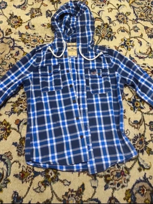 Blårutig skjorta med huva Hollister - Säljer en blårutig skjorta från Hollister med huva och vita snören. Skjortan har två bröstfickor med knappar och klassisk knappstängning framtill. Perfekt för en avslappnad och cool stil.