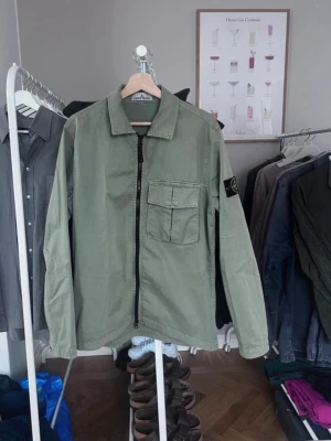 Grön overshirt från Stone Island - Säljer en grön overshirt från Stone Island med klassisk krage, bröstficka med lock och dragkedja framtill. Materialet är robust bomull och har den ikoniska Stone Island patchen på vänster ärm. Perfekt för lager-på-lager och snygg till jeans.