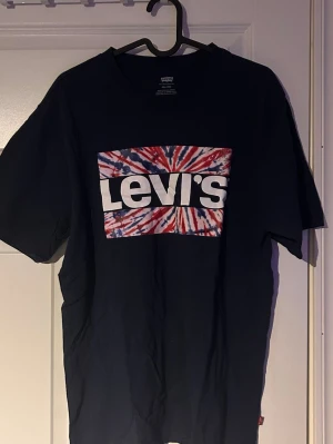  Levi's t-shirt med batiktryck -  t-shirt från Levi's med stort batikmönstrat tryck i rött, blått och vitt på bröstet. Klassisk passform med korta ärmar och rund hals. .