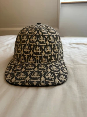 Svartvit keps från Ferregamo - Snygg keps från Ferregamo med svartvitt mönster av logotyper över hela kepsen. Klassisk form med böjd skärm och justerbar rem baktill. Tillverkad i tygmaterial som känns både trendigt och stilrent.