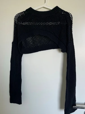 Svart stickad mesh bolero - Trendig svart stickad bolero med meshmönster och långa ärmar. Kort modell som passar perfekt över linnen eller klänningar. Ribbstickad kant vid hals och nederkant för extra detalj. Perfekt för lager på lager och en edgy look.