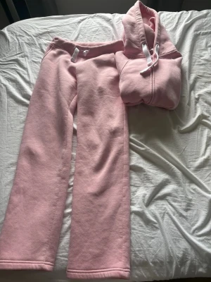 Rosa mjukisset med luvtröja - Mysigt loungewear-set i ljusrosa fleece med matchande byxor och luvtröja. Byxorna har resårmidja med vita dragsnören och raka ben. Luvtröjan har dragkedja och justerbar huva. Perfekt för chill dagar hemma.