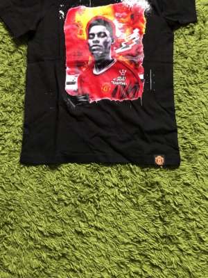 Manchester United svart t-shirt - Snygg svart t-shirt med tryck av en Manchester United-spelare på framsidan och klubbens logga nertill. Motiv i rött, vitt och gult ger en cool kontrast. Klassisk rund hals och korta ärmar. Perfekt för dig som älskar fotboll och vill visa ditt lag.