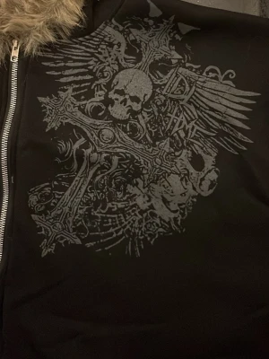 Svart hoodie fuskpäls - Svart hoodie med dragkedja, stor grå dödskalle- och korsdesign på bröstet samt huva fodrad med ljusbrun fuskpäls. Hoodien har en edgy vibe och passar perfekt för dig som gillar streetwear och detaljerade tryck.