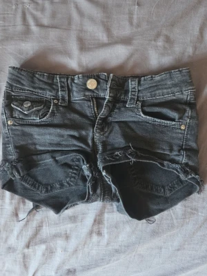 Mörkgrå denimshorts med fransar - Säljer ett par mörkgrå denimshorts med råa fransade benslut och slitna detaljer. Shortsen har fem fickor, bälteshällor och stängs med knapp och dragkedja. Perfekta för sommardagar och festivaler.
