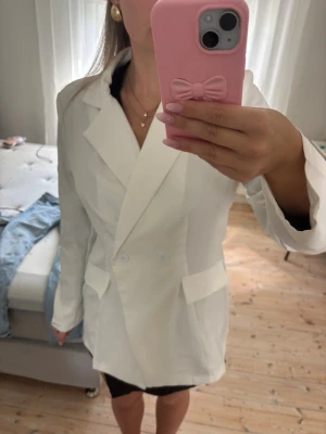 Vit oversized kavaj med dubbelknäppning och figursydd  - Stilren vit kavaj i oversized modell med dubbelknäppning framtill och breda slag. Kavajen har två fickor med lock och långa ärmar. Perfekt för en trendig och clean look. Materialet ser ut att vara en slät bomulls- eller polyestermix.