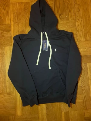 Mörkblå hoodie från Polo Ralph Lauren - Mörkblå hoodie från Polo Ralph Lauren med vit broderad logga på bröstet och vita dragsnören i huvan. Klassisk känguruficka framtill och ribbade muddar vid ärmslut och nederkant. Perfekt för en avslappnad och trendig look.
