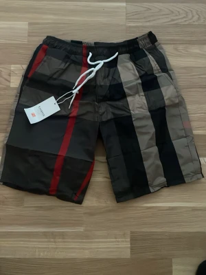 Randiga burberry shorts med snörning - Säljer ett par snygga burberry shorts med breda ränder i svart, beige och rött. Shortsen har elastisk midja med vit snörning och sidofickor. Perfekta för sommaren och har en avslappnad passform.