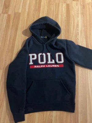 Mörkblå  hoodie från Polo Ralph Lauren - Snygg mörkblå hoodie från Polo Ralph Lauren med stor vit och röd logga på bröstet. Klassisk huva med dragsko och känguruficka framtill. Perfekt för en avslappnad och trendig stil. SKRIV OM DU HAR NÅGON FRÅGA, passar dig som är typ 165-177