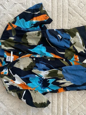 Färgstark kortärmad skjorta från Zara - Säljer en kortärmad skjorta från Zara med ett stort, färgstarkt blommigt mönster i blått, orange, svart, vitt och grönt. Skjortan har klassisk krage och knappar framtill. Perfekt för dig som vill sticka ut med en cool och unik look.