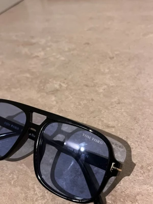 Svarta Tom Ford solglasögon - Snygga solglasögon från Tom Ford med svart båge och blåtonade glas. Klassisk fyrkantig form med diskreta gulddetaljer vid skalmarna. Perfekt accessoar för att lyfta din stil.