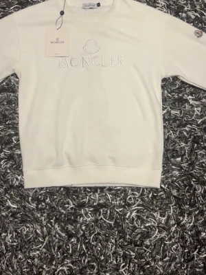 Vit sweatshirt från Moncler - Vit sweatshirt från Moncler med broderad logotyp på bröstet och patch på ärmen. Klassisk rund halsringning och ribbade muddar. Perfekt för en clean och stilren look.