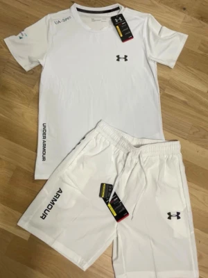Vitt träningsset från Under Armour - Snyggt vitt träningsset från Under Armour med t-shirt och shorts. Båda plaggen har logga och tryck i svart, samt UA-SPIC-detaljer. Materialet är lätt och andas, perfekt för träning eller sport. Elastisk midja på shortsen för skön passform.