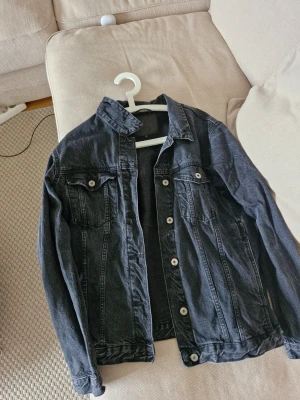 Svart jeansjacka med klassisk look - Säljer en svart jeansjacka med klassisk passform och silverfärgade knappar. Jackan har två bröstfickor med lock och knapp samt sömdetaljer framtill och baktill. Perfekt att slänga över en hoodie eller t-shirt för en avslappnad stil. Står L men passar dig som har m eller s