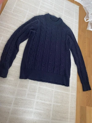 Mörkblå kabelstickad tröja - Mörkblå kabelstickad tröja med rund hals och långa ärmar. Tröjan har breda ribbade muddar vid ärmslut och nederkant. Perfekt för dig som gillar klassisk och stilren look. Materialet känns mjukt och värmande.