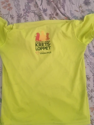 Gul funktions-tshirt Kretsloppet - Neongul funktions-tshirt från Kretsloppet Borås med tryck på bröstet. T-shirten är tillverkad i ett lätt och snabbtorkande material, perfekt för löpning eller träning. Rund hals och korta ärmar. Snygg och sportig design med logga och text.