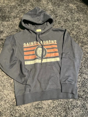 Navy ysl hoodie - Snygg grå hoodie från Saint Laurent med huva och dragsko. Framsidan har ett vintageinspirerat tryck med texten 'Saint Laurent' och ränder i orange och vitt samt ett emblem i mitten. Klassisk känguruficka och ribbade muddar. Perfekt för en avslappnad streetstyle.