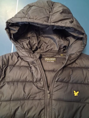Svart pufferjacka från Lyle & Scott - Snygg svart pufferjacka från Lyle & Scott med huva och dragkedja framtill. Jackan har det klassiska gula logomärket på bröstet och två sidofickor. Tillverkad i ett lätt och vadderat material som håller värmen under kalla dagar.