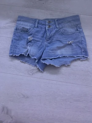 Low waist light washed jeans shorts - Säljer ett par ljusblå denimshorts med fransig kant och slitna detaljer framtill. Shortsen har låg midja, fem fickor och dubbla knappar i linningen. Perfekta för sommaren och ger en avslappnad vibe.