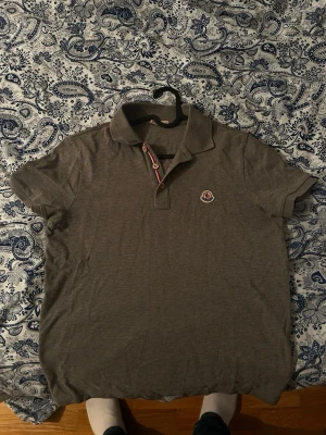 Grå Moncler piké t-shirt - Snygg grå pikétröja från Moncler med klassisk krage och knappslå med rödvit detalj. Tröjan har korta ärmar och en broderad Moncler-logga på bröstet. Tillverkad i mjuk bomull som passar perfekt till en avslappnad stil. Kvitto finns även.