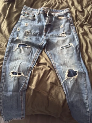 Blå slitna jeans med hål - Säljer ett par ljusblå jeans med rejält slitna detaljer och stora hål på både fram- och baksida. Jeansen har klassisk femficksmodell och raka ben. Perfekta för dig som gillar en edgy och avslappnad stil.