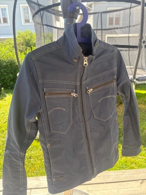 Mörkblå jeansjacka från G-Star RAW - Snygg mörkblå jeansjacka från G-Star RAW med hög krage, dragkedja framtill och två bröstfickor med dragkedja. Kontrastsömmar ger en cool look och jackan har justerbar midja baktill. Perfekt för dig som vill ha en stilren och modern jeansjacka.