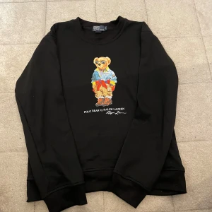 Svart tröja Polo Bear Ralph Lauren M - Svart långärmad tröja från Polo Ralph Lauren med ikoniskt Polo Bear-tryck framtill. Tröjan har rund halsringning och normal passform. Materialet är mjukt och skönt, perfekt för en chill och stilren look.