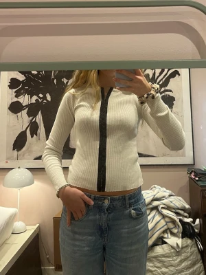 Vit ribbad cardigan med svarta detaljer - Säljer en vit ribbad cardigan med lång ärm och svarta detaljer längs knäppningen framtill. Tröjan har en tight passform och rund halsringning. Perfekt att styla med jeans eller kjol för en clean look.