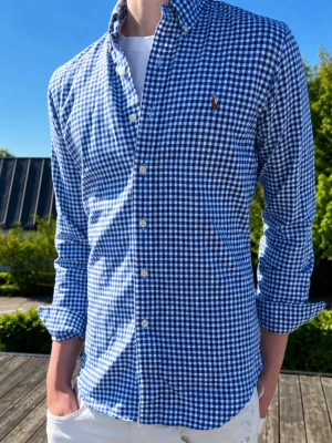 Blårutig skjorta från Ralph Lauren - (MODDELEN ÄR 185 OCH VÄGER 75kg)Klassisk blårutig skjorta från Ralph Lauren med button-down krage och vita knappar. Skjortan har lång ärm och den ikoniska broderade loggan på bröstet. Perfekt till jeans eller chinos för en clean och stilren look.