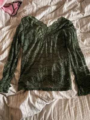 Grön spetsblus med v-ringning - Snygg mörkgrön långärmad topp i genombruten spets med v-ringad hals. Blusen har ett romantiskt mönster och är lätt transparent, perfekt att styla med ett linne under. Materialet är mjukt och följsamt, vilket ger en feminin siluett. Storleken är XL men skulle säga att den sitter mer M/L 