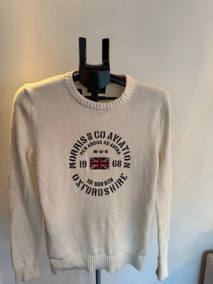 Vit stickad tröja Morris Oxfordshire - Vit stickad tröja från Morris med rund halsringning och tryck framtill med brittisk flagga och texten 'Morris & Co Aviation Oxfordshire'. Tröjan har långa ärmar och ribbade muddar vid ärmslut och nederkant. Perfekt för en avslappnad och stilren look. Stor i storlek.