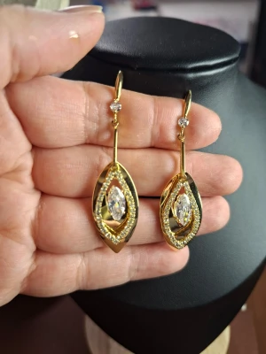 Guldiga hängande örhängen med stenar - Snygga guldiga örhängen med hängande design och glittrande stenar i mitten. Örhängena har en oval form med dubbla lager och små gnistrande detaljer runt om. Perfekta för dig som gillar statement-smycken och vill sticka ut.