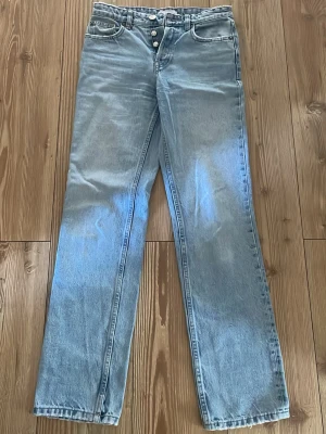 Ljusblå raka jeans med knappar - Säljer ett par ljusblå jeans med raka ben och hög midja. Jeansen har synliga silverfärgade knappar i gylfen och klassiska fem fickor. Tillverkade i denim med en avslappnad passform, perfekta för en chill och trendig look.