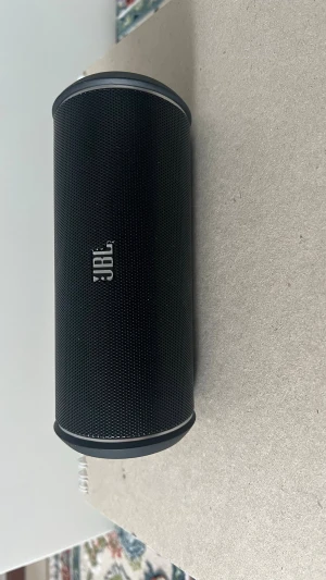 JBL svart trådlös högtalare - Snygg svart trådlös högtalare från JBL med cylinderformad design och robust metallgaller. Högtalaren har tydliga knappar på sidan för enkel styrning av musik och samtal. Perfekt för att ta med överallt tack vare sin kompakta storlek och stilrena look.