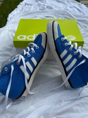 Blå Adidas sneakers med vita ränder (42) - Snygga blå sneakers från Adidas med klassiska vita ränder på sidorna och vita snören. Skorna har en högre modell och är tillverkade i textil med gummisula. Perfekta för dig som vill ha en sportig och stilren look.