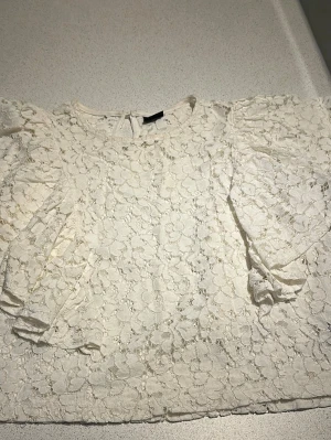Vit spetsblus från Gina Tricot - Superfin vit blus i spets från Gina Tricot med blommigt mönster och rund halsringning. Blusen har korta ärmar med lätt puff och knäppning bak i nacken. Perfekt att styla med jeans eller kjol för en trendig look.
