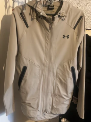 Beige vindjacka från Under Armour - (FINNS FULLT MED KLÄDER INNE SKRIV PRIVAT DET DU BEHÖVER )Snygg beige vindjacka från Under Armour med svarta detaljer och dragkedja framtill. Jackan har huva med dragsko och två fickor med svarta dragkedjor. Perfekt för blåsiga dagar och sportiga outfits.Har använt dd. ett par gånger säljs för det passar inte min storlek , den är såklart äkta under Armour påsen följer med priset kan diskuteras för allt
