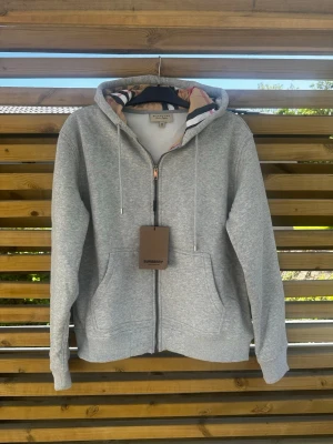 Grå hoodie från Burberry med dragkedja - Snygg grå hoodie från Burberry med dragkedja framtill och klassiskt rutigt foder i huvan. Tröjan har två stora fickor, ribbade muddar och justerbar dragsko i huvan. Perfekt för en avslappnad och stilren look.